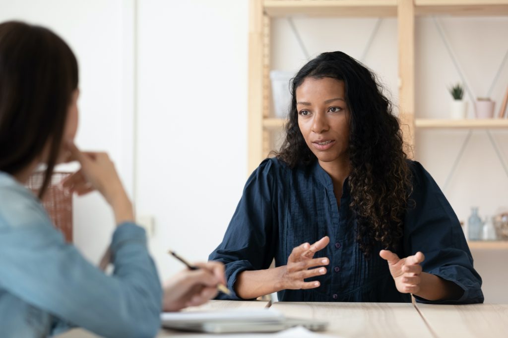 young woman with long dark hair speaking to another woman across a table